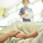 a hospital visitor's hand holds a patient's hand in bed of a hospital ward. In the blurred background a young nurse is chatting to the ward sister about the patient's care.