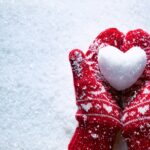 Female hands in knitted mittens with snowy heart against snow background. Love, winter and Valentines day romantic creative concept with copy space for text